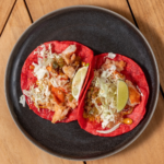 Lobster tacos on red tortillas with cabbage and lime wedges on a black plate