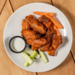 Smoked chicken wings with a cup of dipping sauce, carrot and celery sticks on a white plate